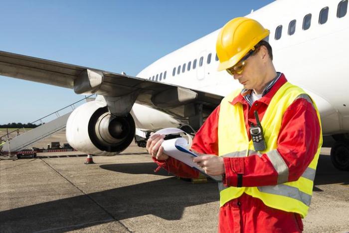 Maintenance technician in a yellow vest and hard hat inspecting an airplane on the tarmac, holding a clipboard and notepad in hand